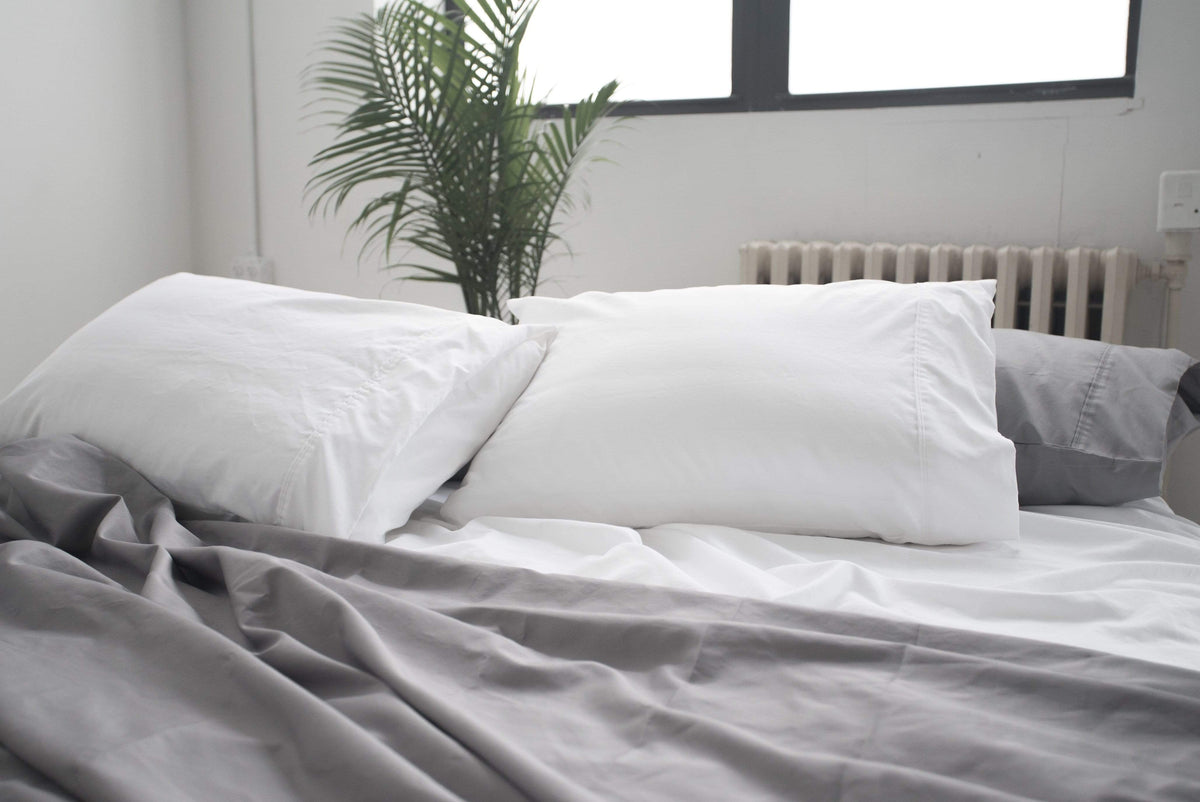 Unmade bed with white pillows and gray sheets in a bright bedroom.