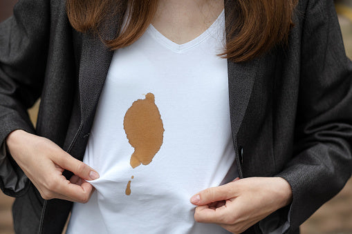 A person wearing a dark grey blazer over a white v-neck t-shirt with a large, dark brown coffee or food stain on the center of the chest.