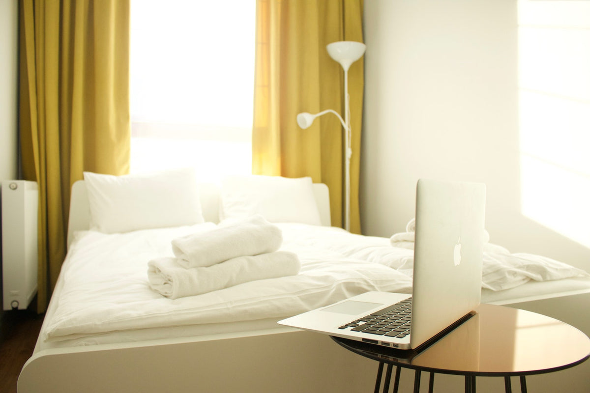 Bright bedroom with neatly made bed, folded towels, and laptop on side table
