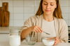 A woman in a kitchen measuring white powder with a spoon from a glass bowl into a smaller bowl.