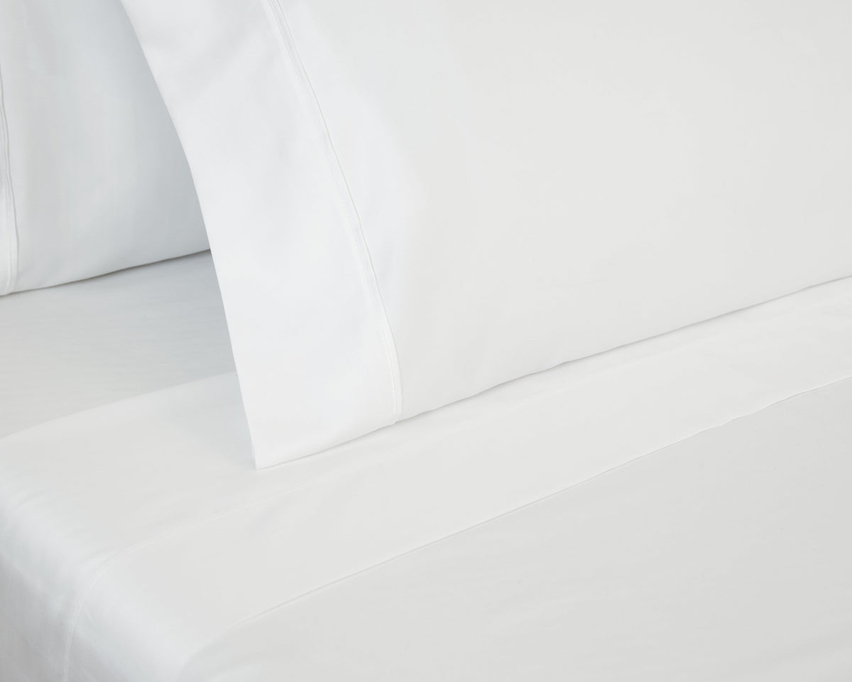 A clean, minimalist close-up of a bed styled with crisp white pillowcases and a matching flat sheet neatly tucked in, showcasing a smooth and hotel-quality fabric finish.