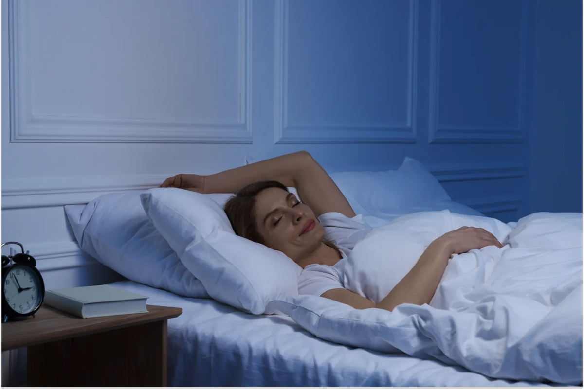 A woman sleeping soundly in a bed with crisp white linens, under cool blue night lighting, with a book and a black alarm clock on a wooden bedside table.