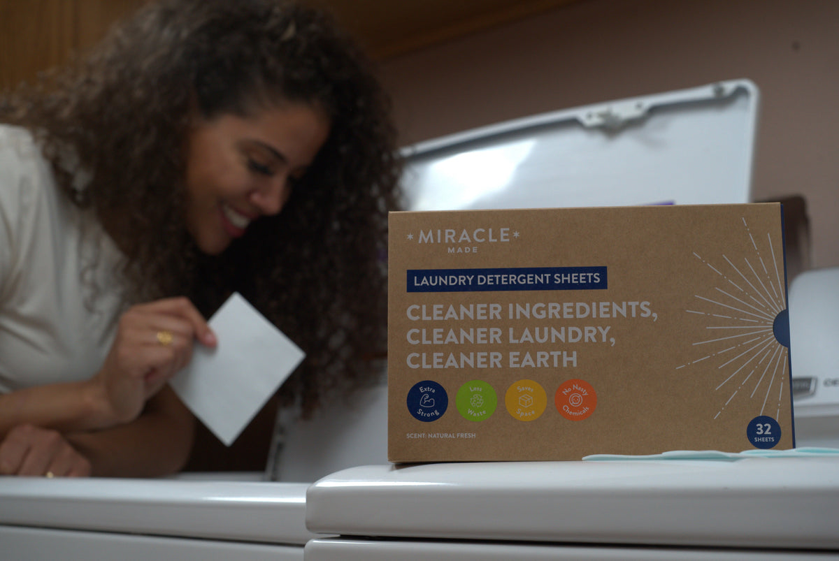 A box of Miracle Made Laundry Detergent Sheets sitting on a white washing machine, with a smiling woman in the background holding a single white detergent sheet.