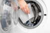 A close-up of a hand placing a thin, white rectangular laundry detergent sheet into the drum of a front-loading washing machine.
