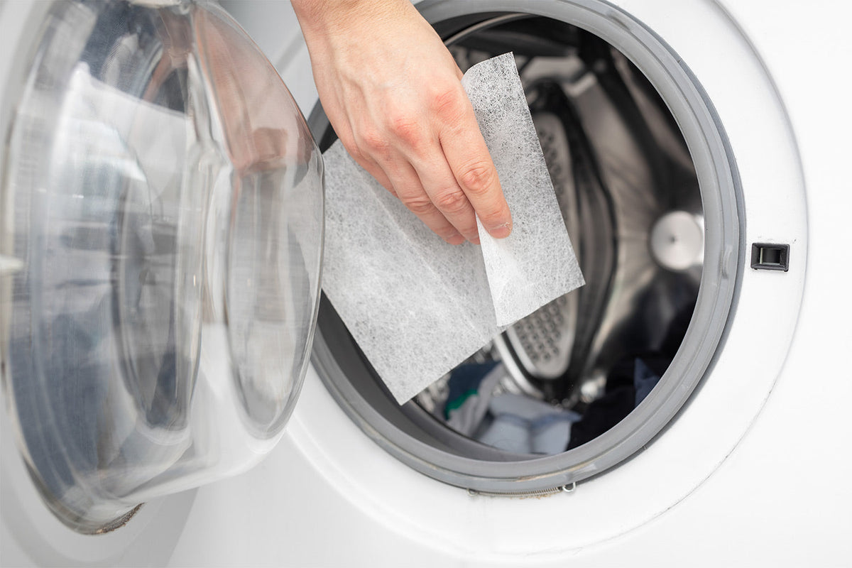 A close-up of a hand placing a thin, white rectangular laundry detergent sheet into the drum of a front-loading washing machine.