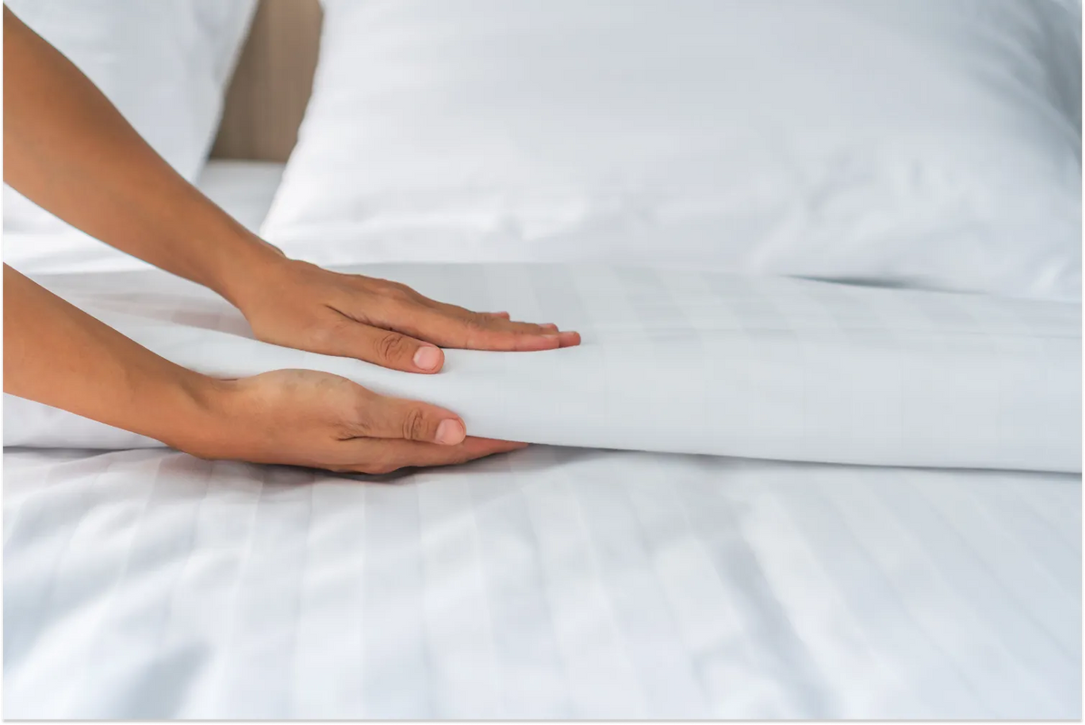 Close-up of a person's hands smoothing a crisp, white striped linen sheet on a neatly made bed.