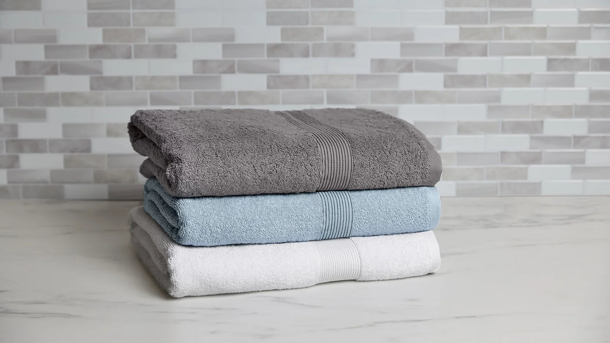A stack of three neatly folded plush bath towels in Stone grey, Sky blue, and White, displayed on a marble countertop against a light grey tiled backsplash.