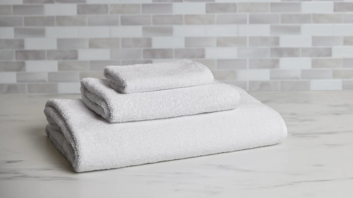 A stack of three neatly folded white plush towels in varying sizes, including a large bath towel, a hand towel, and a washcloth, displayed on a white marble surface against a light grey tiled wall.