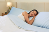 A woman sleeping peacefully on her side in a bed styled with light blue Miracle Made sheets and pillowcases under a white comforter.