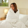 A woman sitting upright in bed with her eyes closed, smiling peacefully while wrapped in a thick, white Miracle Made cooling comforter.