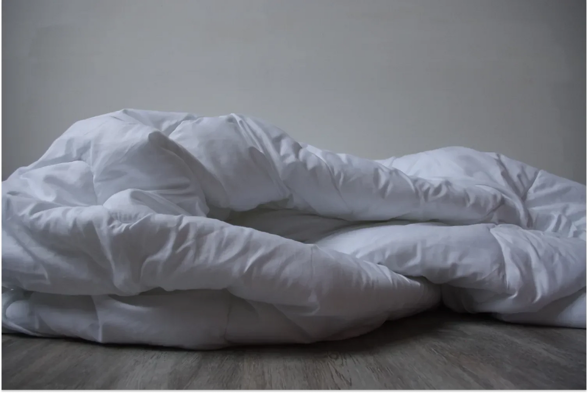 A large, fluffy white comforter or duvet piled in soft, voluminous folds on a light wood floor against a plain grey wall, highlighting a crisp percale texture.