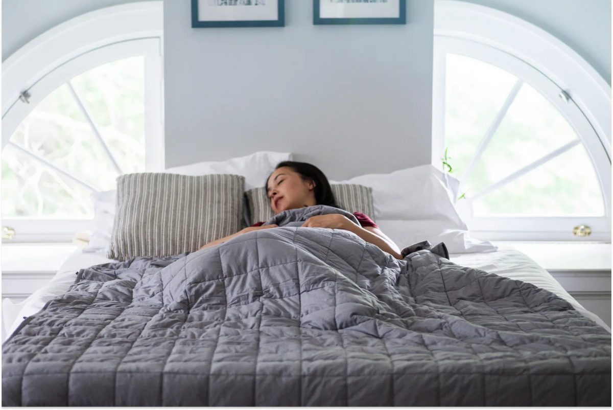 A woman sleeping comfortably in a bed positioned between two arched windows, covered by a grey quilted weighted blanket and resting on striped pillows.