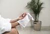 A close-up of a person in a white bathrobe drying their hands with a small, fluffy white cotton hand towel in a bright room with a potted palm tree in the background.