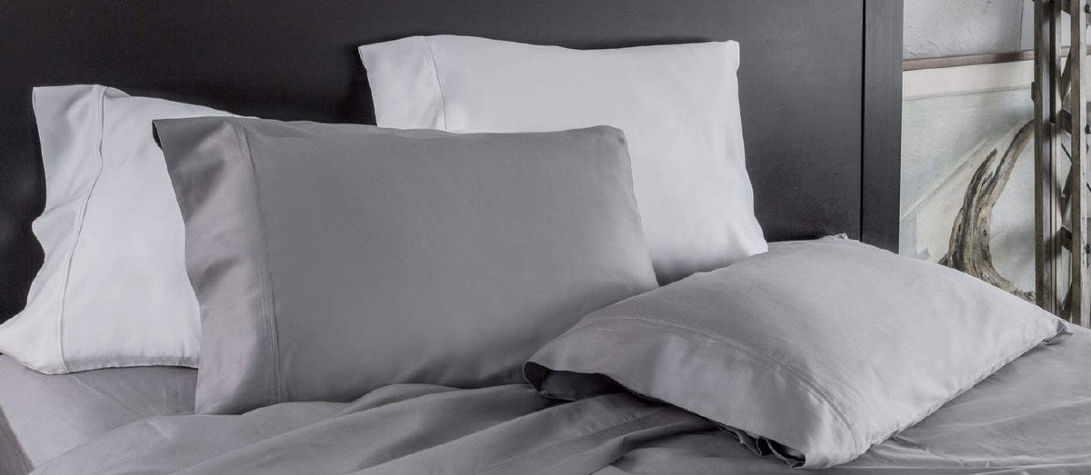 A close-up of a bed styled with a mix of light grey and white pillowcases and matching grey sheets against a dark headboard.
