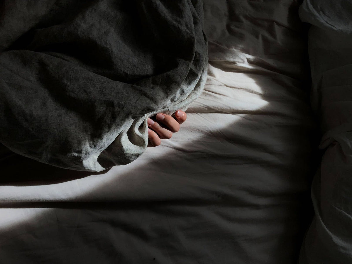 A dramatic high-contrast shot of a hand clutching the edge of a dark grey duvet on a bed with light-colored wrinkled sheets, illuminated by a sliver of morning sunlight.