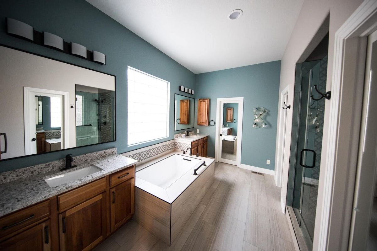 A modern master bathroom featuring a large rectangular drop-in bathtub, a double vanity with granite countertops and wooden cabinets, and slate blue walls.