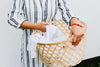 A woman in a navy and white striped button-down shirt holding a woven bamboo laundry basket filled with white linens against a plain white background.