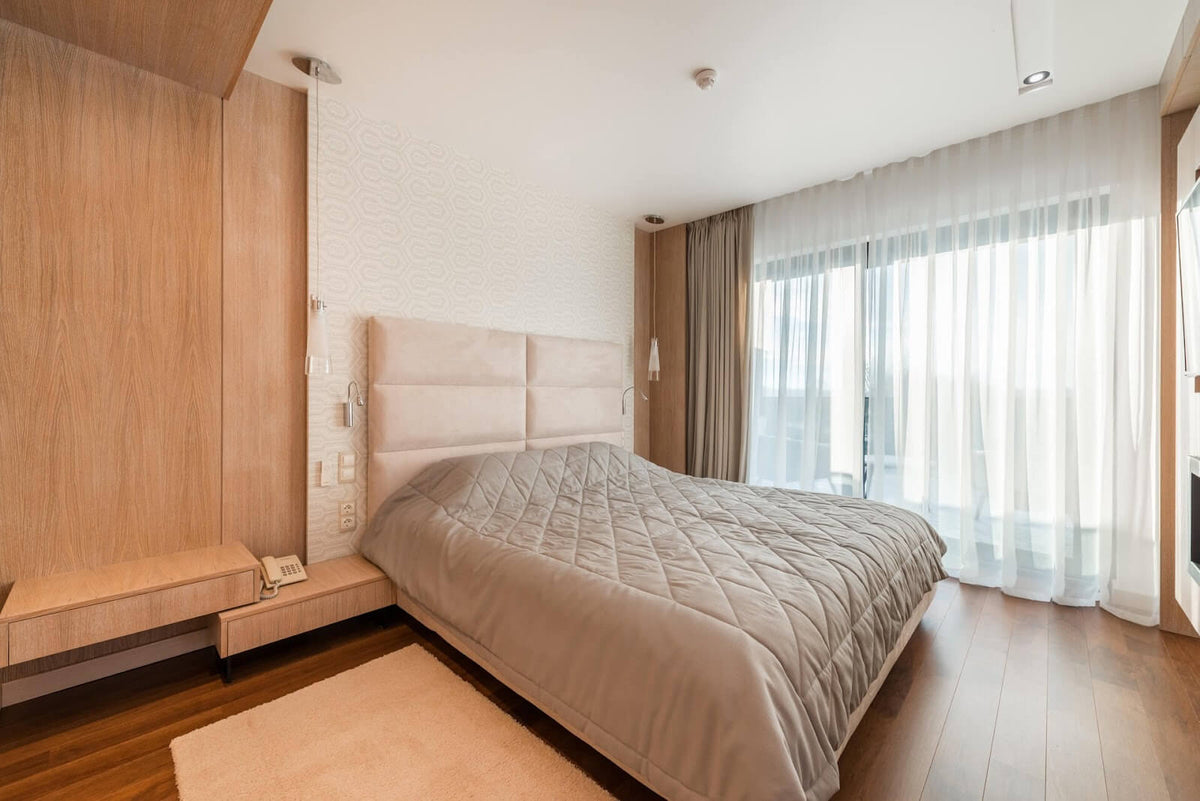 A bright, modern bedroom featuring a large bed with a tan quilted comforter, a high beige headboard, light wood wall paneling, and sheer white floor-to-ceiling curtains.