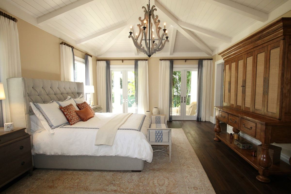 Elegant bedroom with a large bed, white bedding, chandelier, wooden furniture, and tall windows letting in natural light.
