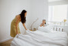 A woman in a yellow dress making a bed with white waffle-weave bedding, while a young girl rests her chin on her hands atop a stack of white pillows.