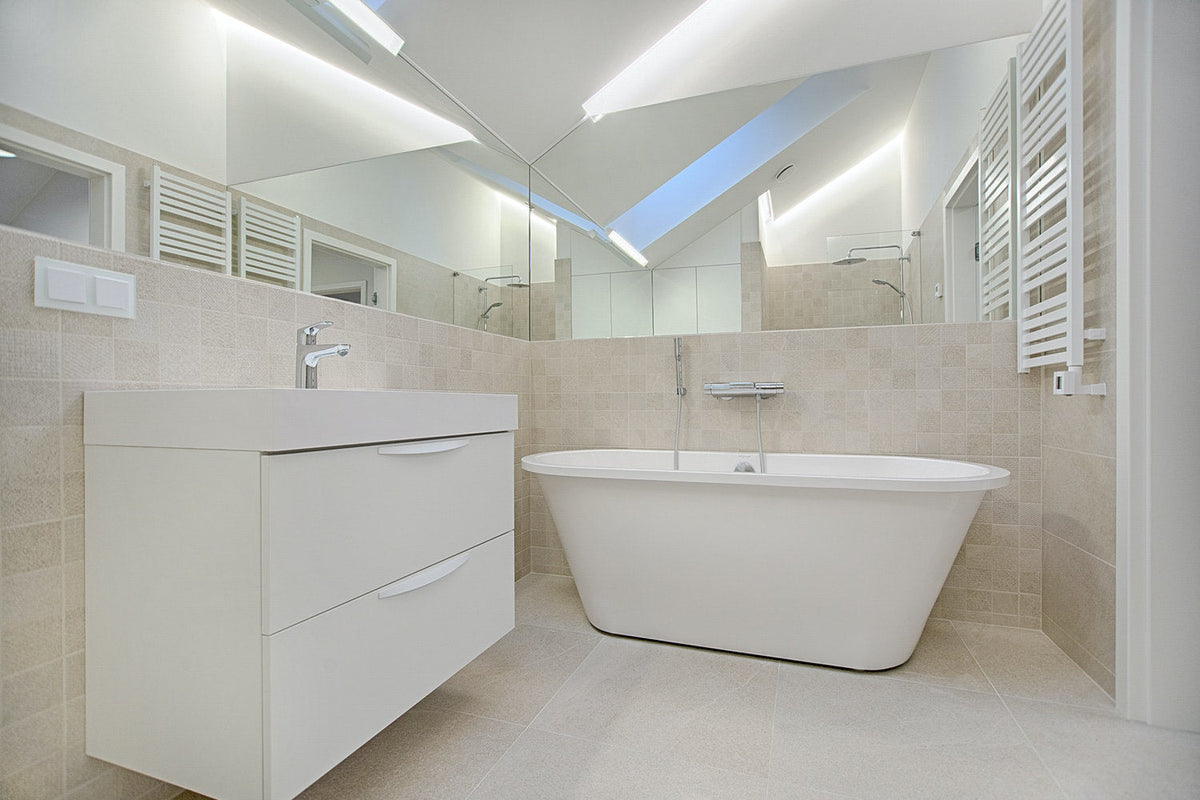 A minimalist, ultra-modern bathroom featuring a white freestanding soaking tub, a sleek white floating vanity with two drawers, and neutral beige wall tiles under bright, recessed lighting.