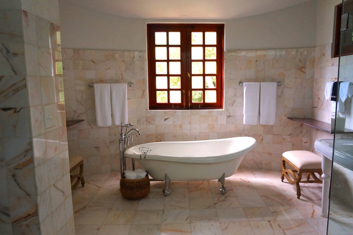 A luxury marble-tiled bathroom featuring a white clawfoot bathtub, white towels on wall-mounted racks, and a large wooden window that lets in natural light.