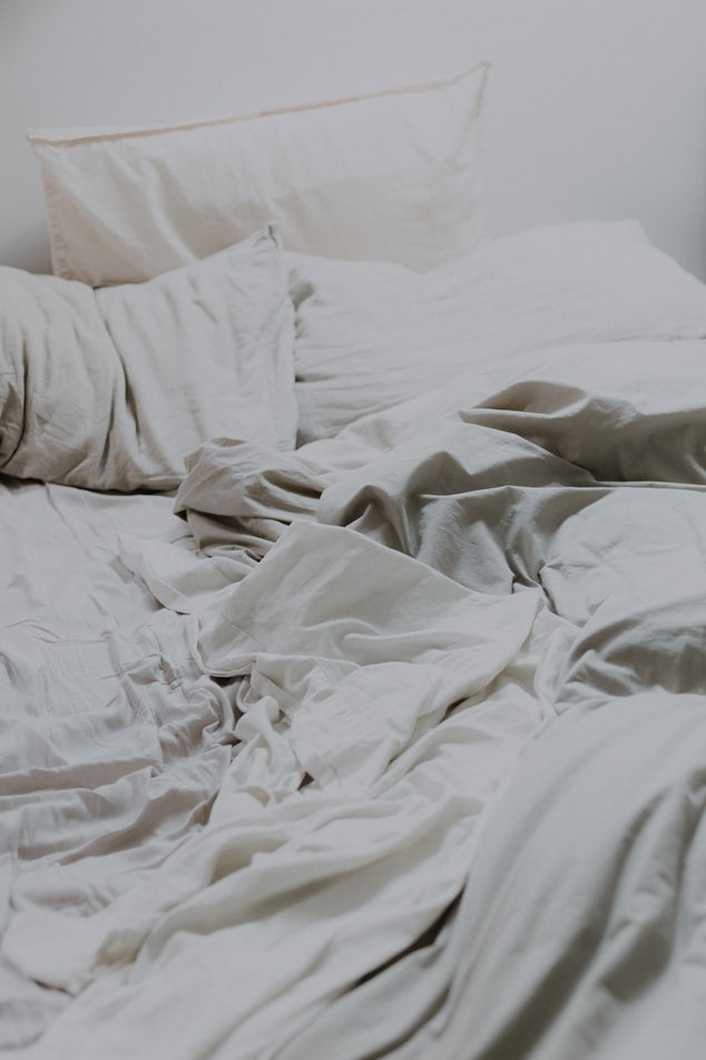 An unmade bed with rumpled light grey or off-white sheets and multiple matching pillows, creating a soft, lived-in aesthetic.