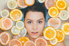 A top-down portrait of a woman submerged in water, surrounded by floating slices of lemons, oranges, and grapefruits to illustrate a refreshing citrus-infused spa bath.