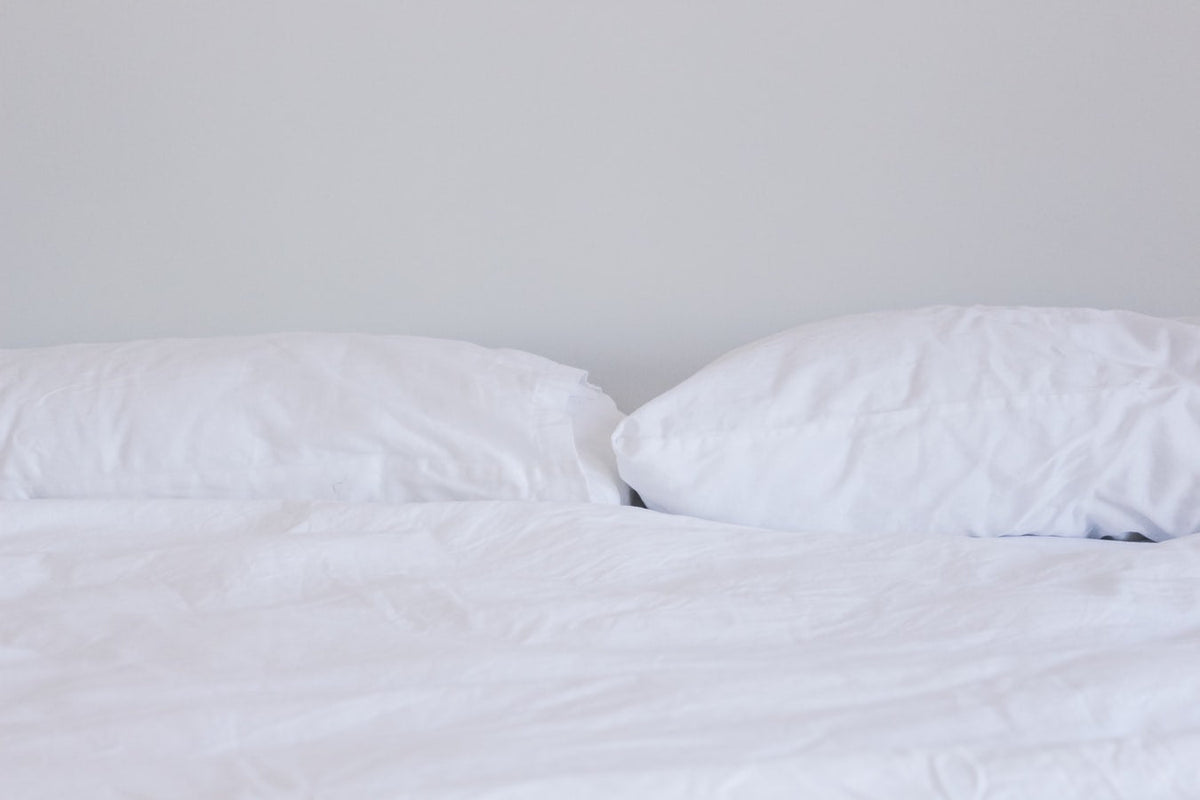 A bright, minimalist bed featuring two slightly rumpled white pillows resting on a white flat sheet against a plain light grey wall.