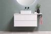A minimalist white floating bathroom vanity with a vessel sink and black hardware, featuring a small potted plant and a pink hand towel hanging from a black rack against a sage green wall.