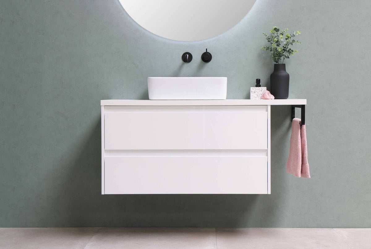 A minimalist white floating bathroom vanity with a vessel sink and black hardware, featuring a small potted plant and a pink hand towel hanging from a black rack against a sage green wall.