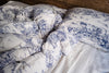 A bed styled with a white and blue toile de Jouy patterned duvet cover and matching pillowcase, featuring classic scenic illustrations on a rumpled white sheet.