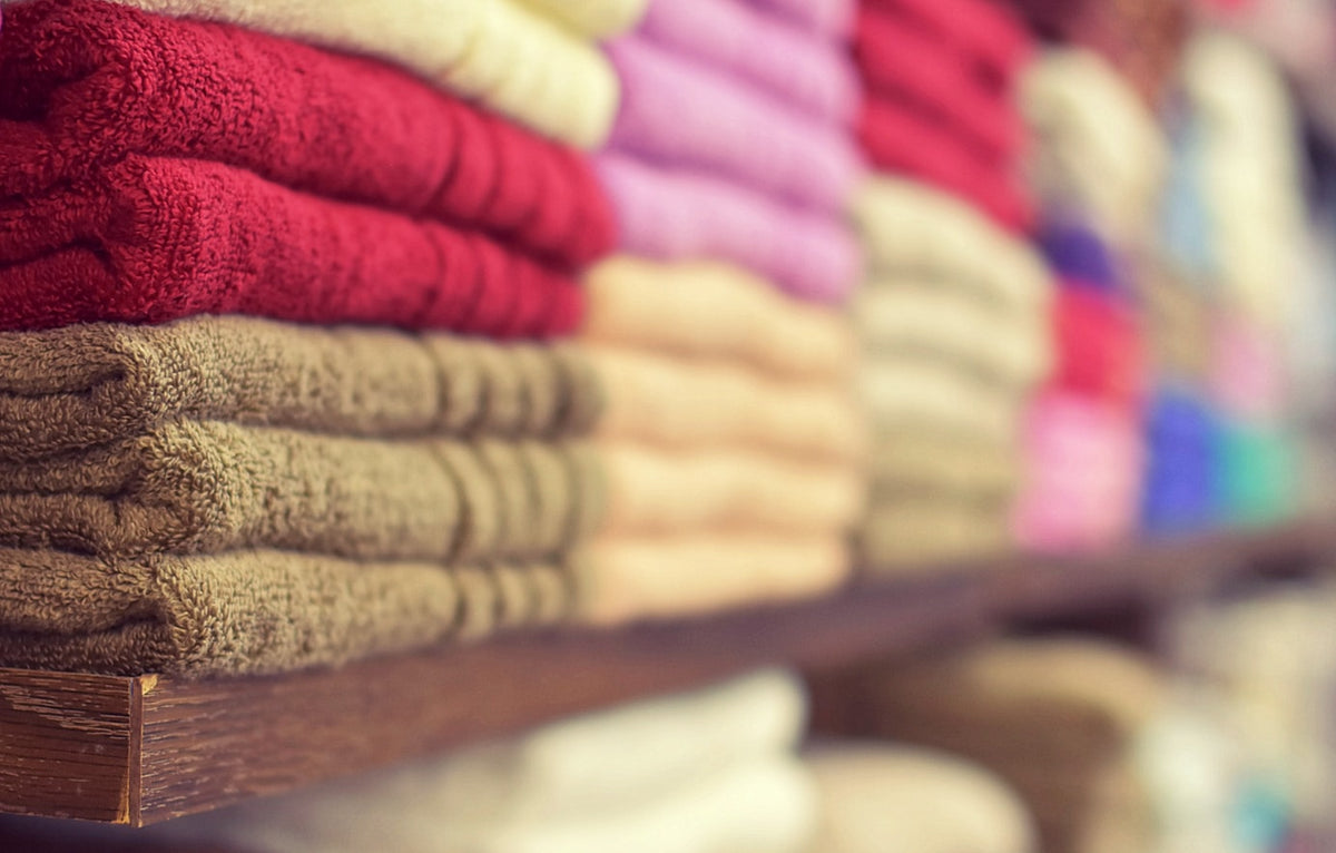 A side view of multiple stacks of colorful, plush cotton towels in shades of red, pink, cream, and tan, neatly arranged on wooden shelving to show variety and texture.