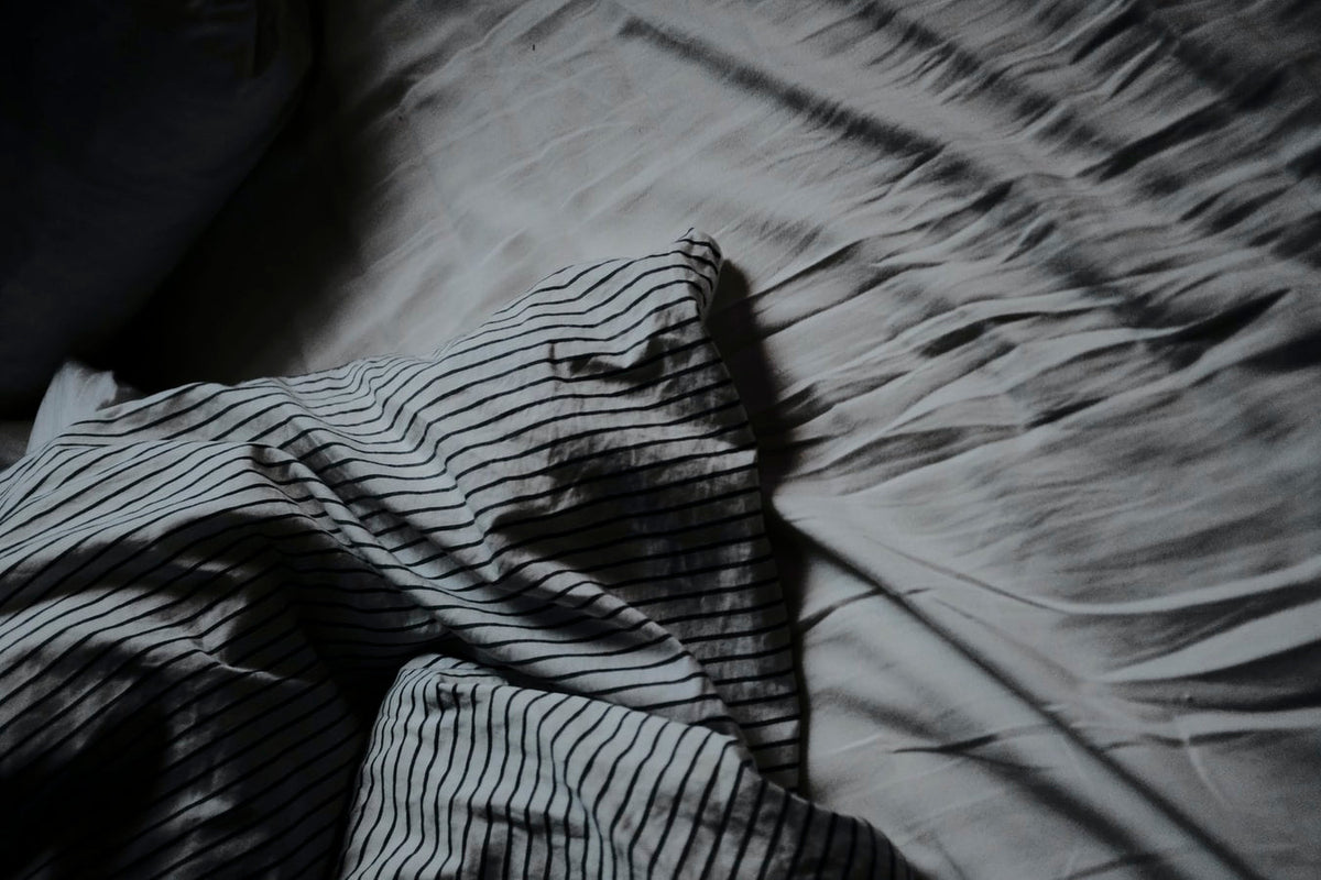 A dramatic high-contrast shot of a bed with rumpled light-colored sheets and a navy and white striped pillowcase, partially obscured by deep shadows.