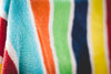 A close-up, abstract view of a vibrant multi-colored beach towel featuring vertical stripes in red, white, turquoise, orange, green, navy blue, and yellow.