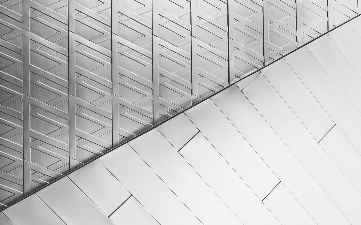 A black and white architectural photograph of a building's exterior, featuring a sharp diagonal line separating a repetitive triangular geometric pattern from smooth, flat panels.