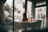 A woman with long blonde hair seen from behind, relaxing in a white freestanding bathtub and holding a glass of water while looking out large windows at a desert landscape with massive boulders.