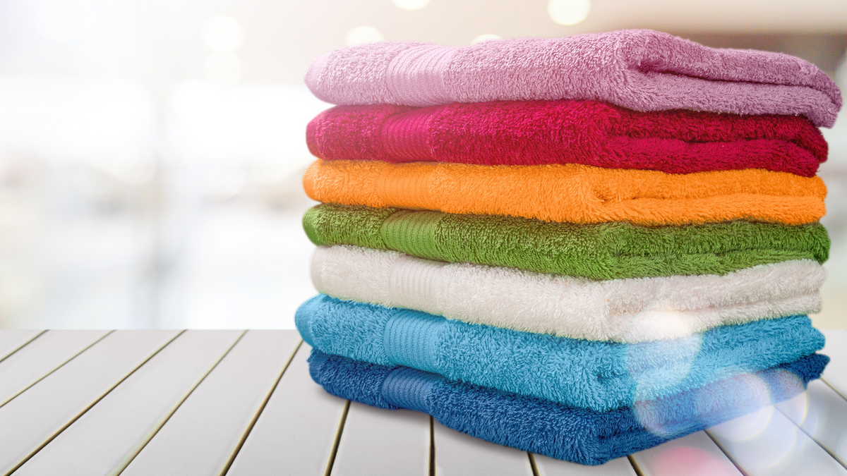 A vibrant stack of seven plush, folded cotton towels in a rainbow of colors—pink, red, orange, green, white, light blue, and navy-resting on a white slatted surface.