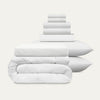 Stack of white bedding including a duvet, comforter, and pillows on a light gray background