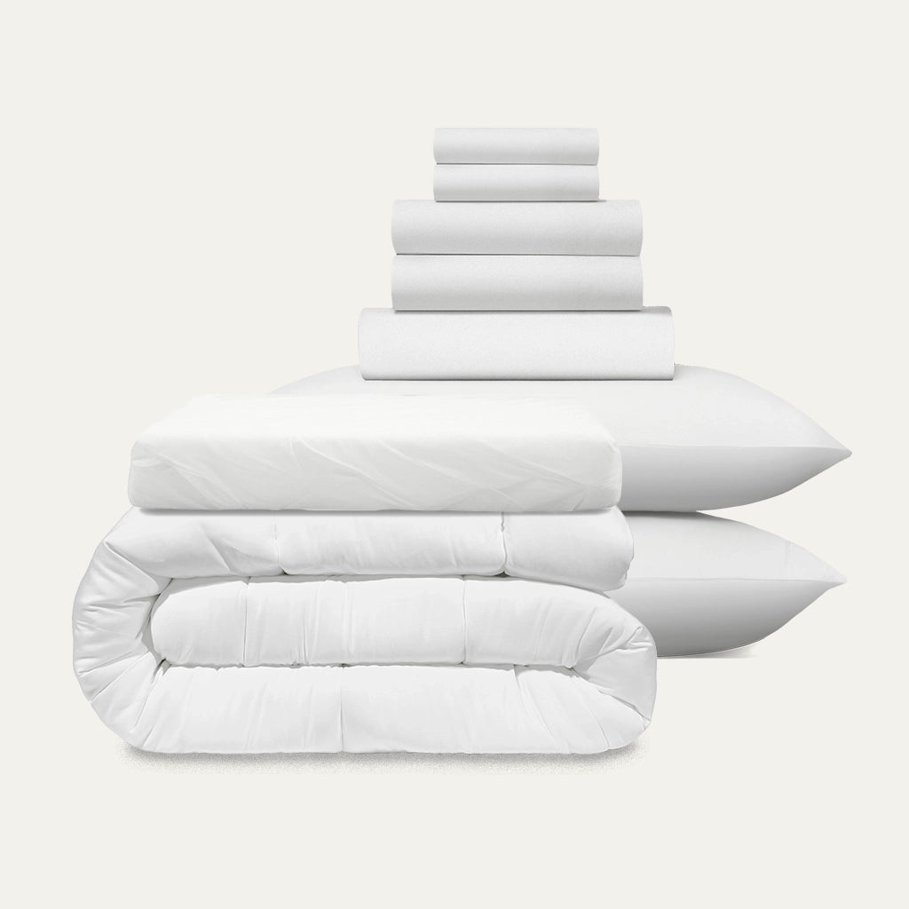 Stack of white bedding including a duvet, comforter, and pillows on a light gray background