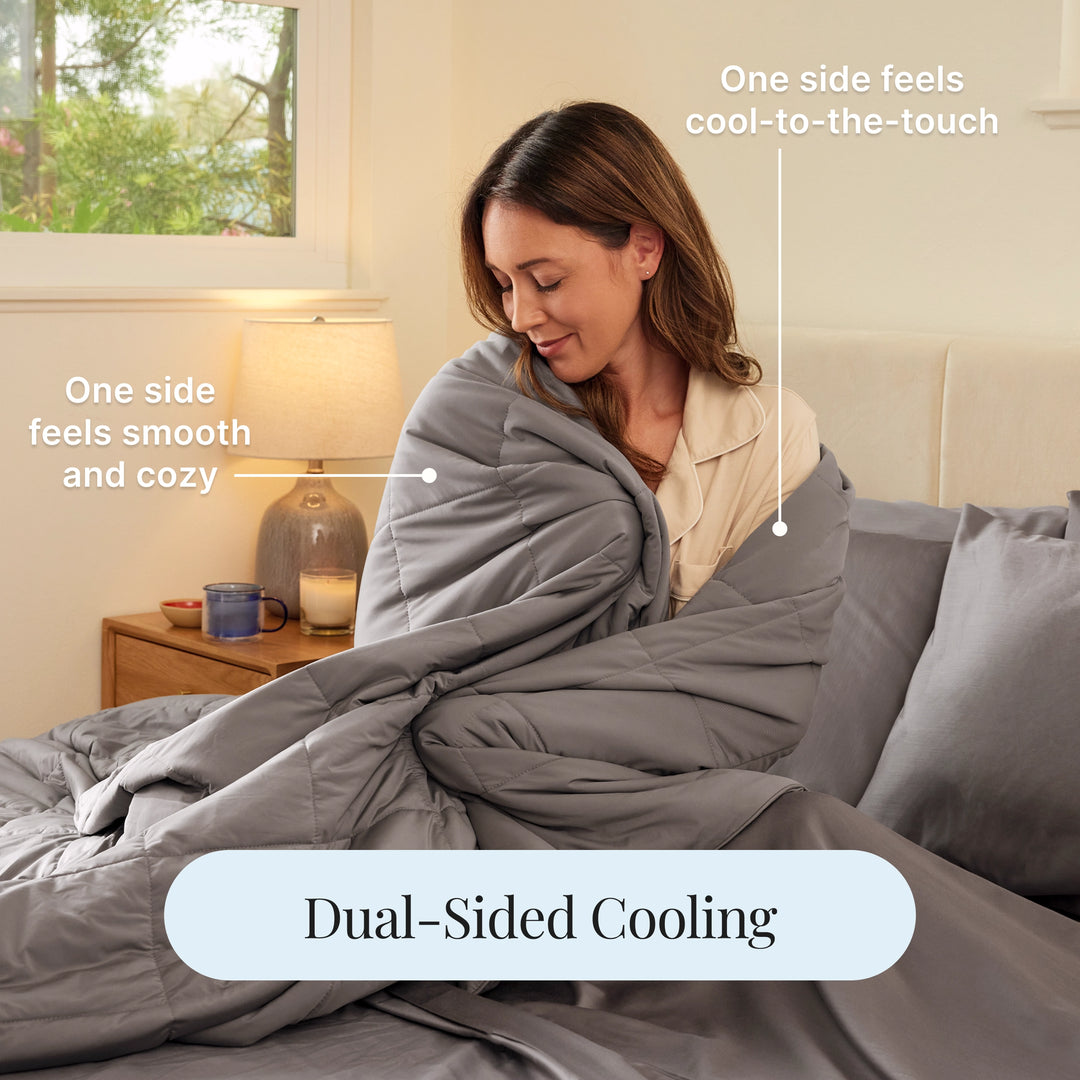 Woman wrapped in a gray dual-sided cooling blanket in a bedroom setting.