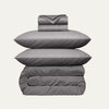 Stack of gray bedding including a duvet, two pillows, and a folded blanket on a light gray background.