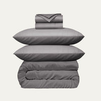 Stack of gray bedding including a duvet, two pillows, and a folded blanket on a light gray background.