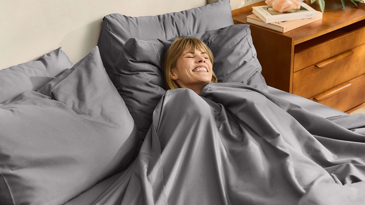 Woman lying in bed with gray bedding in a bedroom setting.