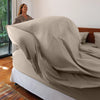 Woman standing next to a bed with a fitted sheet partially covering it in a room with a window and plants.