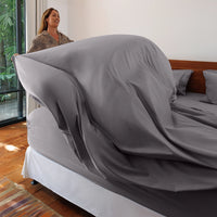 Woman standing next to a bed with a fitted sheet partially covering it in a room with a window and plants.