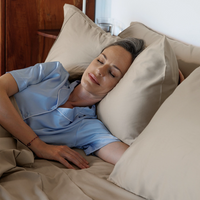 Woman in a blue shirt lying in bed with beige pillows and bedding.