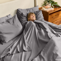 Person lying in bed under gray bedding with a wooden nightstand and books in the background.