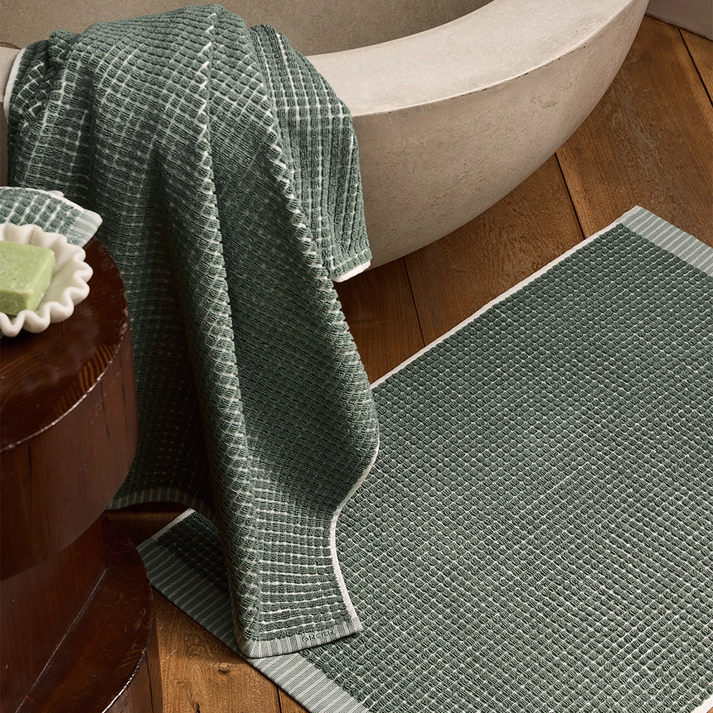 Green textured bath mat in front of a bathtub with a towel draped over the edge.